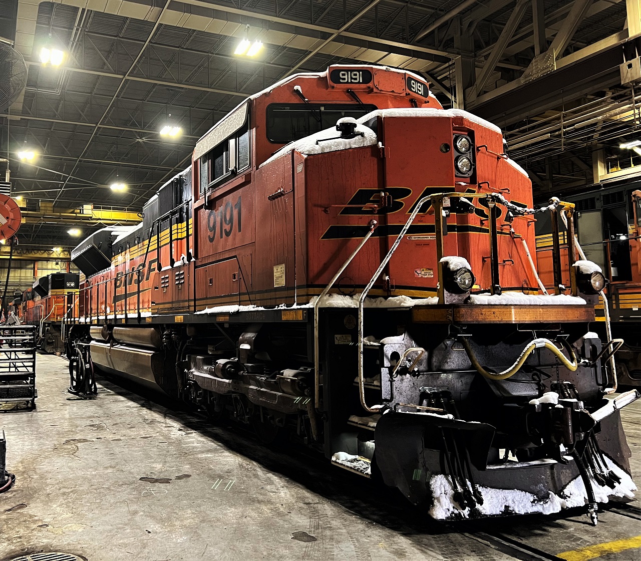 The image depicts a bright orange BNSF locomotive stationed inside an industrial train maintenance facility. The locomotive is well-lit by the overhead lights, with visible signs of snow or frost on its front. The setting features a high-ceilinged space with industrial equipment in the background.