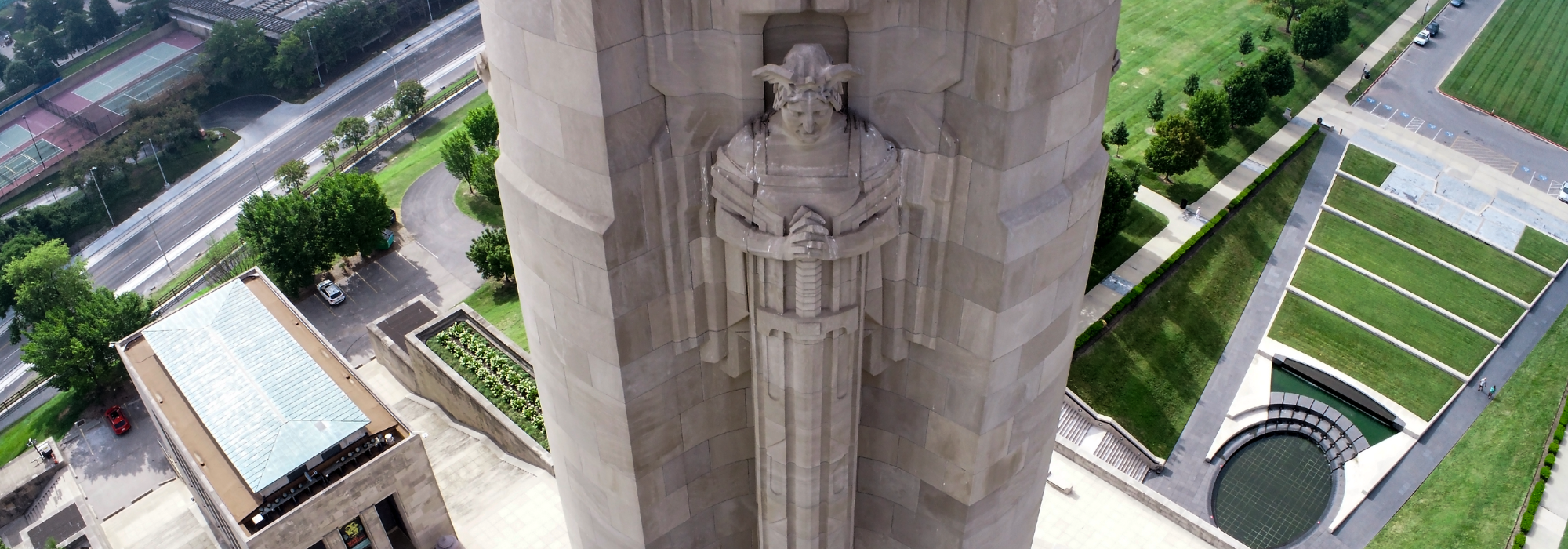 An aerial view of the Liberty Memorial Tower in Kansas City, showcasing the intricate stonework of the monument and the surrounding landscape including a reflecting pool and green terraces.