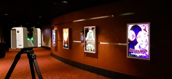 A hallway in an AMC Theater in San Diego with movie posters displayed on the wall. A laser scanner is set up on a tripod in the foreground, capturing dimensional data from the area. The carpet is red/orange, the ceiling is black, and the walls are curved and painted in warm tones.