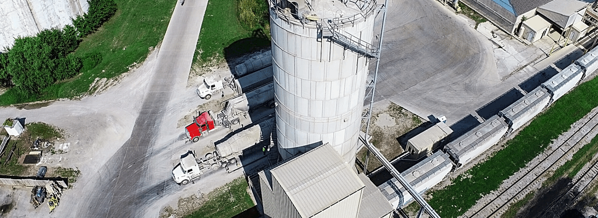 An aerial view of an industrial facility featuring a large cylindrical storage silo, several cement trucks parked nearby, and a train with cargo containers on adjacent railroad tracks.