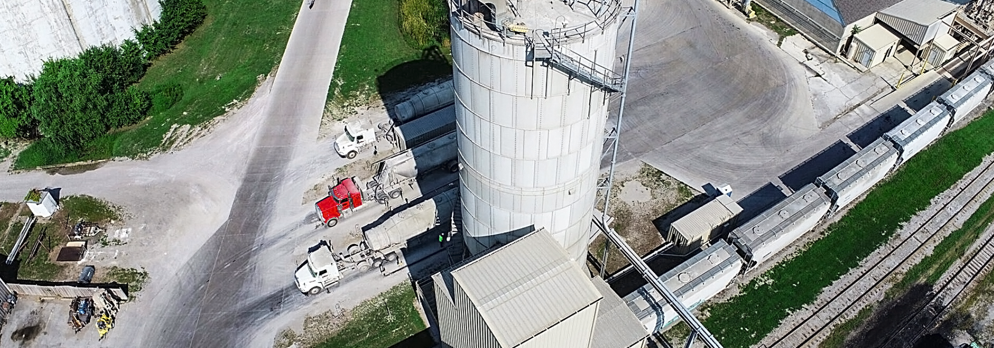 An aerial view of an industrial facility featuring a large cylindrical storage silo, several cement trucks parked nearby, and a train with cargo containers on adjacent railroad tracks.