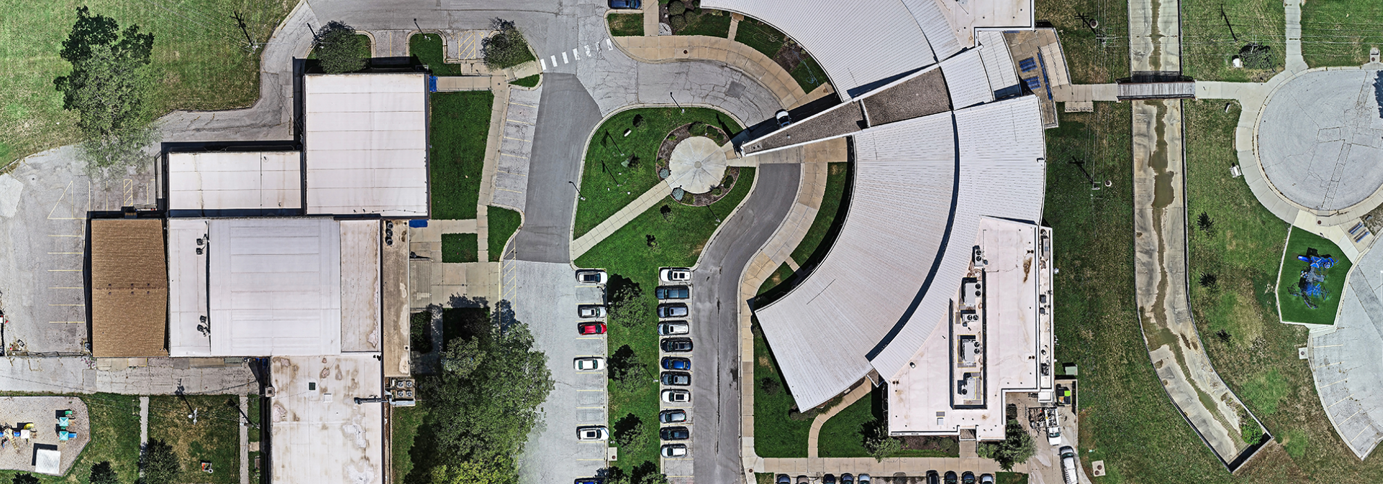 A top-down aerial view of a circular building complex with surrounding parking lots, green spaces, pathways, and adjacent structures.