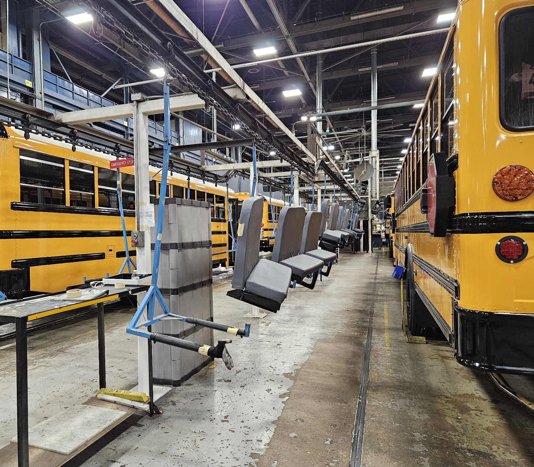 View looking along a pair of assembly lines at a bus factory. Two rows of yellow busses are visible on each side of the image. A single row of gray bench seats hang waiting to be installed.