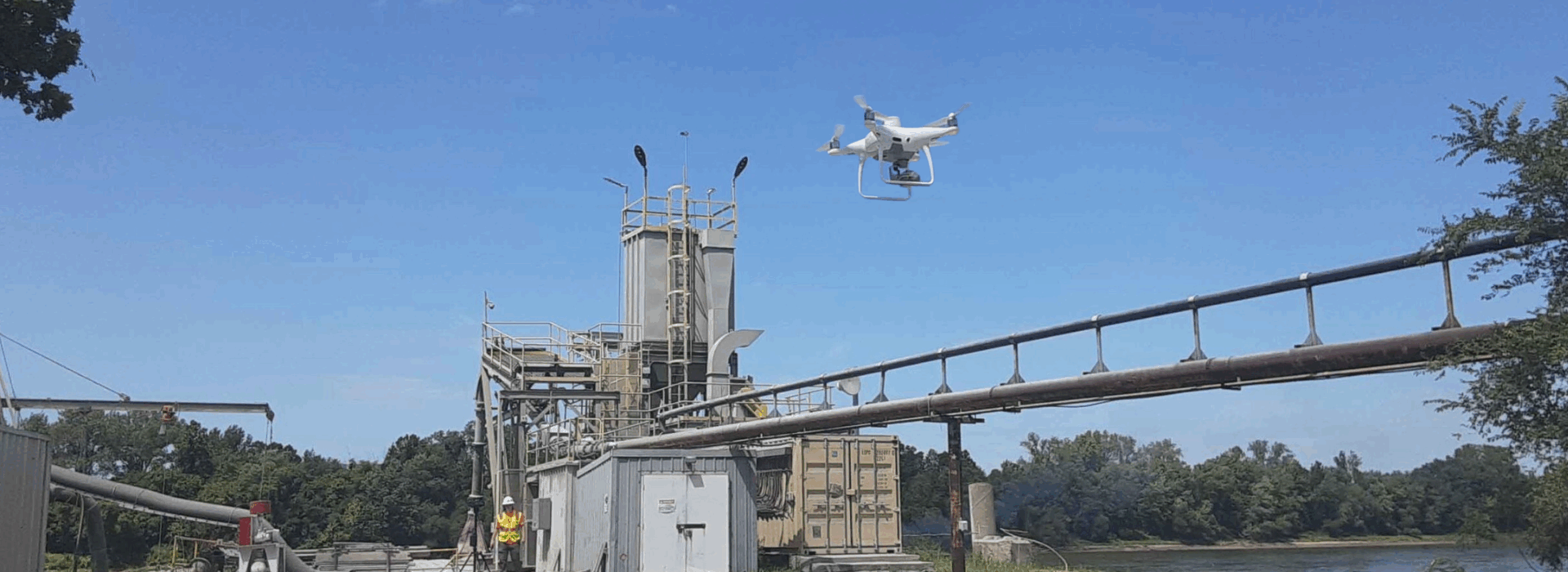 A white drone hovers in the sky above an industrial facility near a river, with metal structures, pipelines, and a worker in a safety vest visible below. The drone is equipped with a camera for aerial inspection and data collection.
