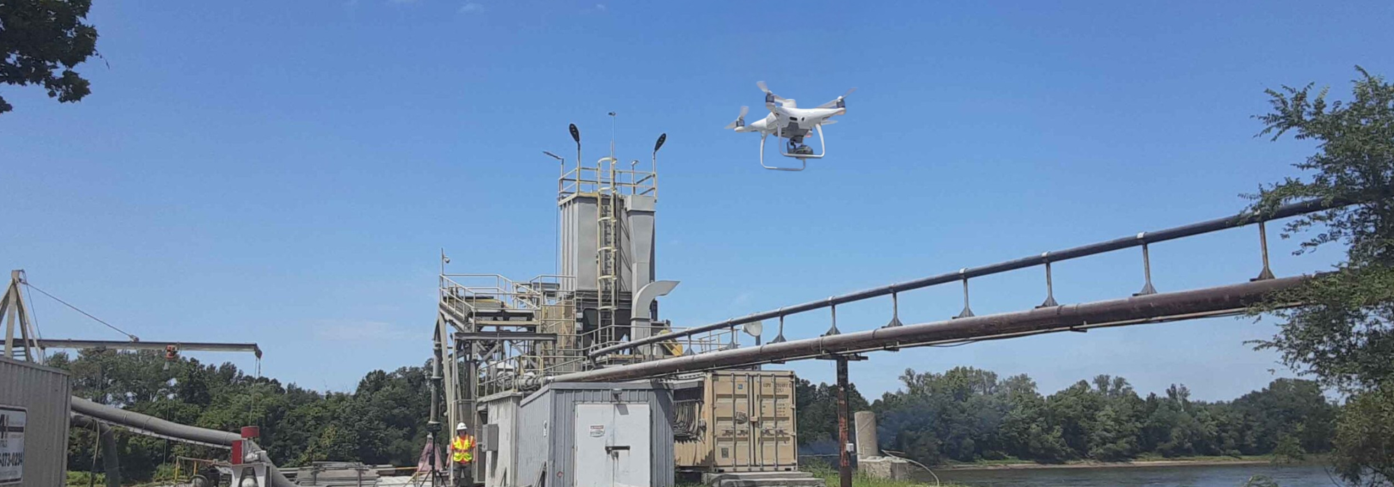 A white drone hovers in the sky above an industrial facility near a river, with metal structures, pipelines, and a worker in a safety vest visible below. The drone is equipped with a camera for aerial inspection and data collection.