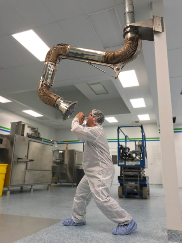 A technician in a white cleanroom suit playfully speaks into a large industrial ventilation pipe in a high-tech scanning facility.